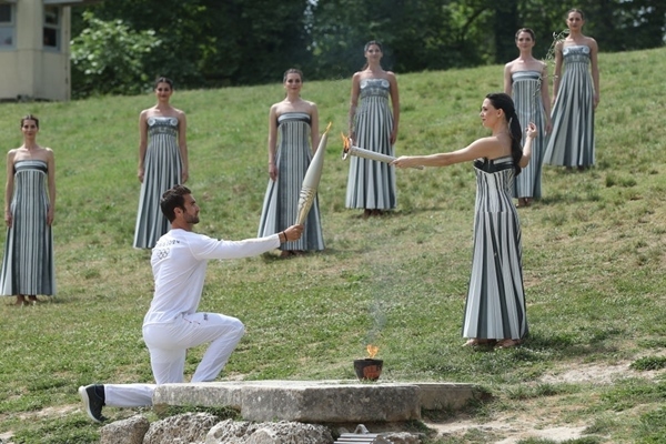 扮演最高女祭司的演員瑪麗·米娜（前右）點(diǎn)燃第一棒火炬手——希臘運(yùn)動(dòng)員、東京奧運(yùn)會(huì)男子賽艇項(xiàng)目金牌得主斯特凡諾斯·恩圖斯科斯手中的火炬。新華社記者 趙丁喆 攝